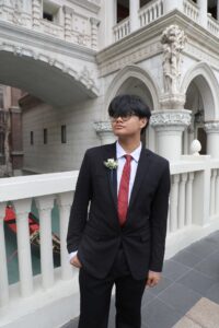 A young person looks off to the distance wearing a formal suit and bright red tie and white boutonniere standing in front of a Venetian building and bridge