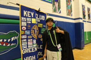 A young person wearing a green and yellow ribbon lei holds a thumbs up standing in front of a Key Club banner